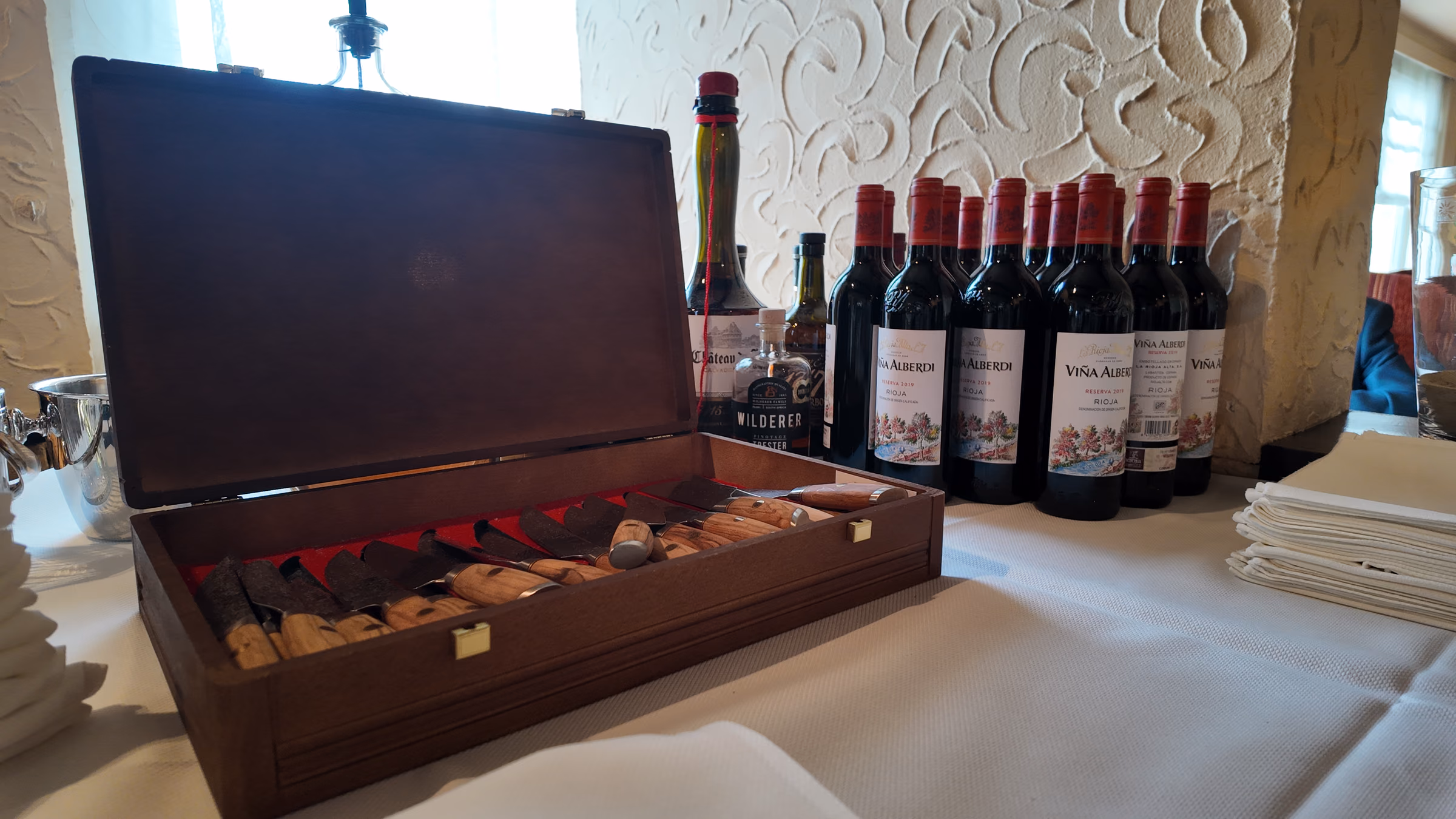 Open wooden case of steak knives on a white tablecloth, with a row of red wine bottles behind