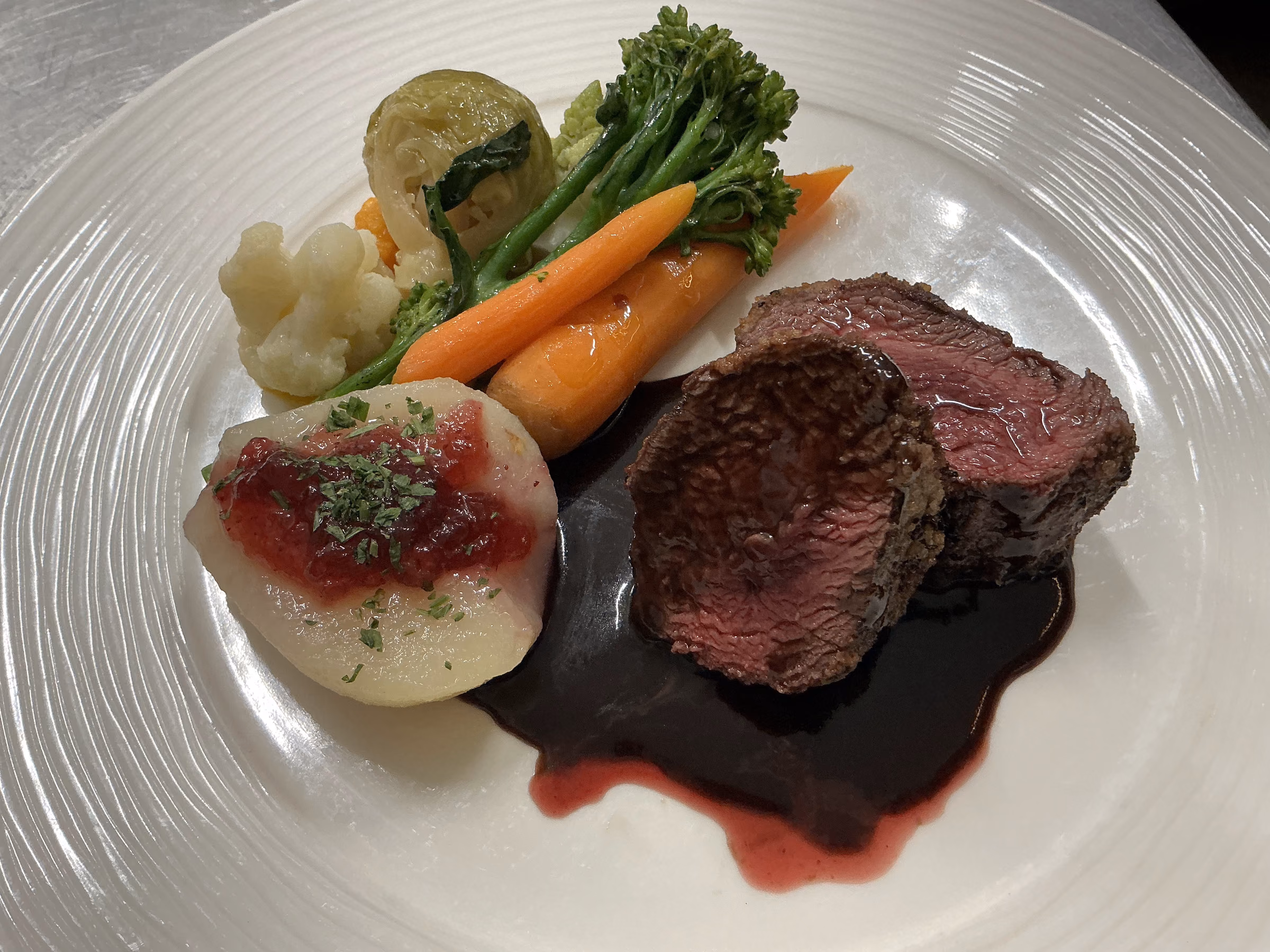 Zwei rosa gebratene Medaillons vom Hirschrücken in dunkler Rotweinjus, daneben eine pochierte Birne mit Preiselbeeren, Broccolini, Babykarotten, Blumenkohl und Rosenkohl
