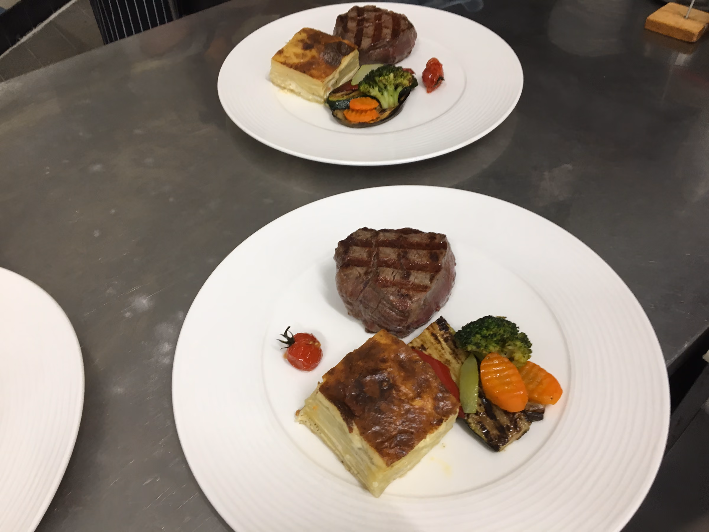 Drei Teller auf dem Anrichtepass, je ein gegrilltes Rinderfiletsteak mit Kartoffelgratin, Grillgemüse und Kirschtomate