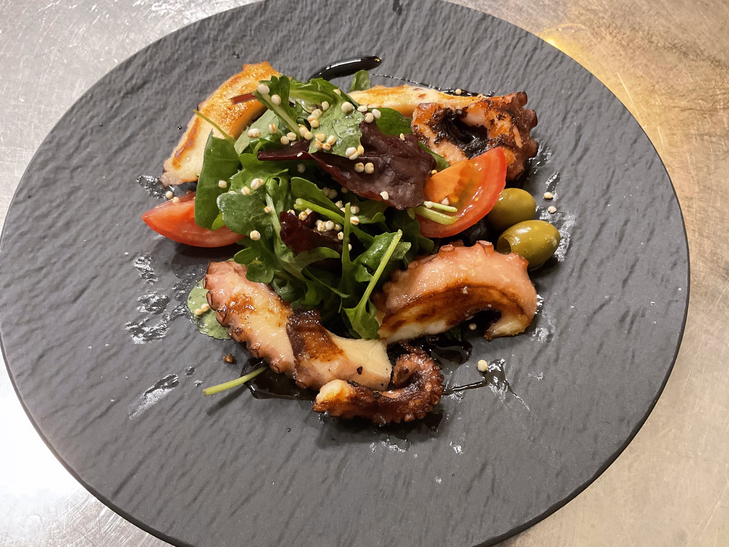 Gegrillte Oktopusarme auf schwarzer Schieferplatte, dazu Rucola, Blattsalat, Tomatenspalten, grüne Oliven, Gerstengraupen und Balsamicoreduktion