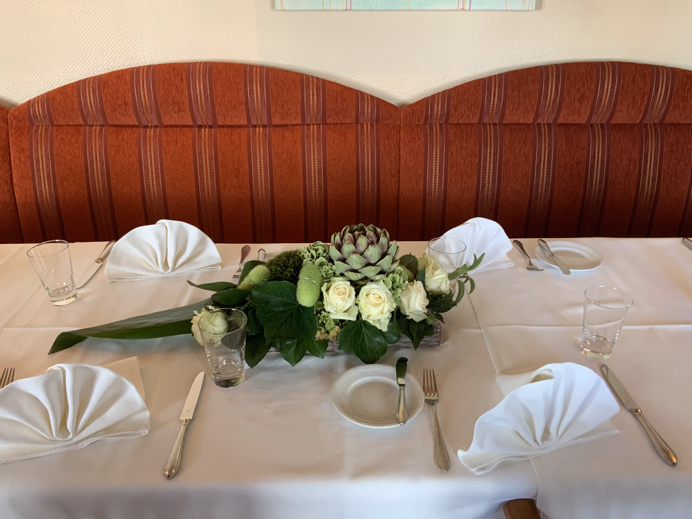 Lange eingedeckte Tafel bei Tageslicht vor der rot gestreiften Polsterbank, mit Serviettenfächern, Gläsern und einem Gesteck aus weißen Rosen, Hortensien und einer Artischocke