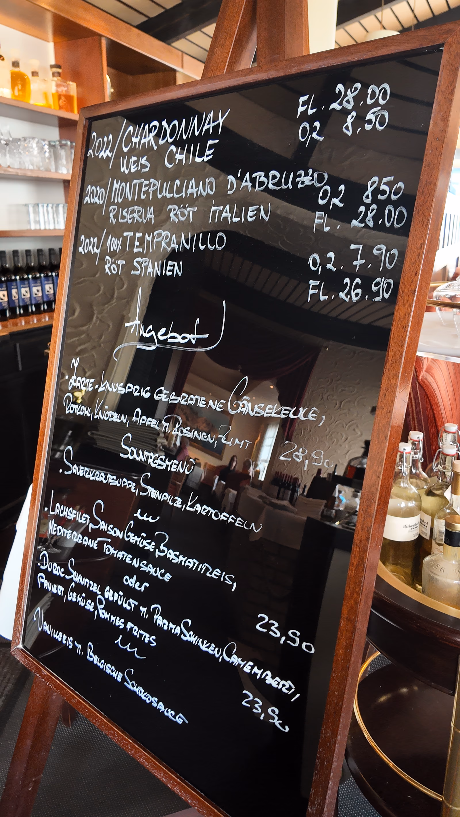 Handwritten board on a wooden easel showing the dishes of the day and the wines by the glass, with the bar shelf to the left